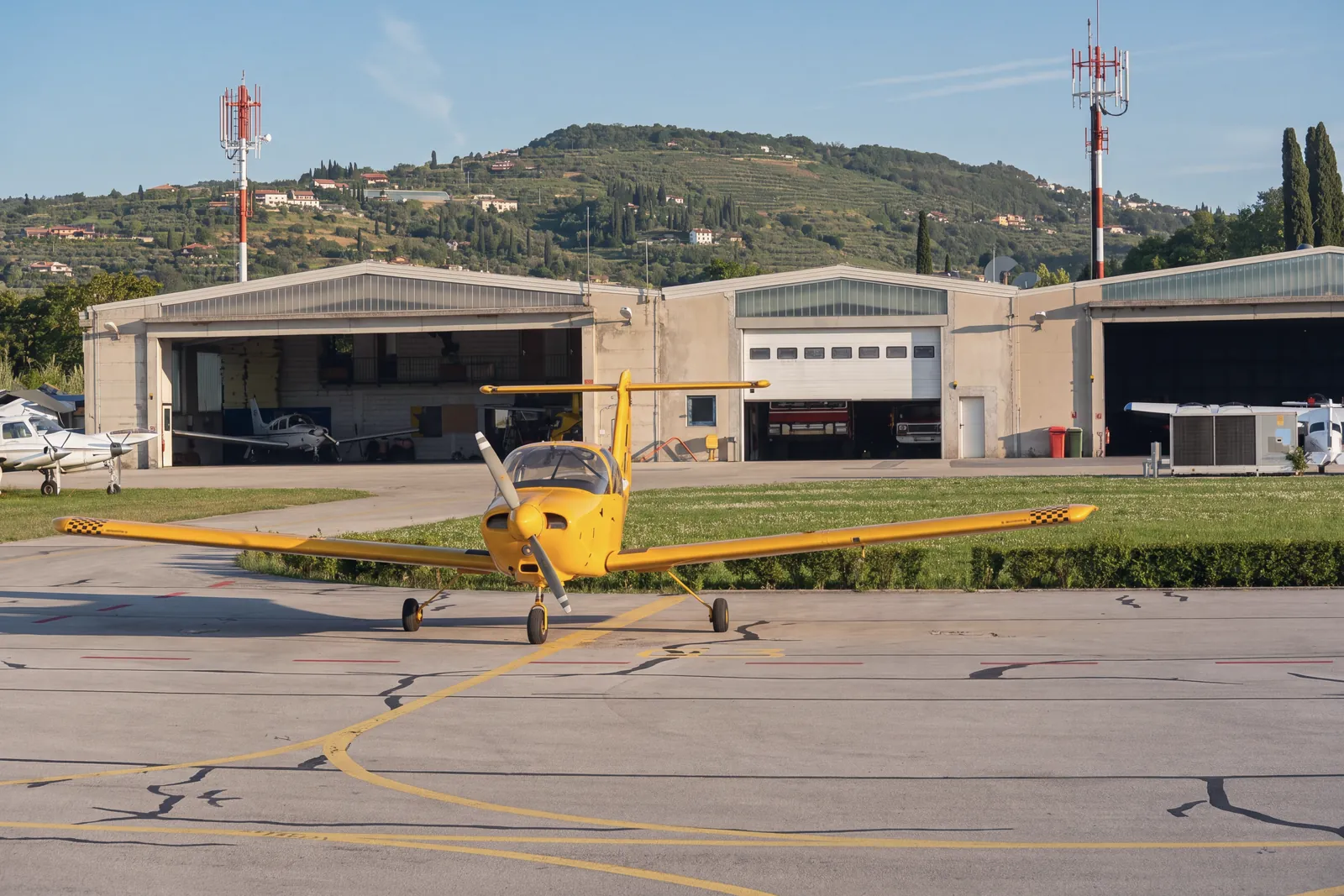A small yellow propeller aircraft parked on the apron of Portorož airport, in front of the hangars; on the left other small aircraft, two red-and-white masts, cypresses and, behind, a terraced hillside with scattered houses. Morning light.