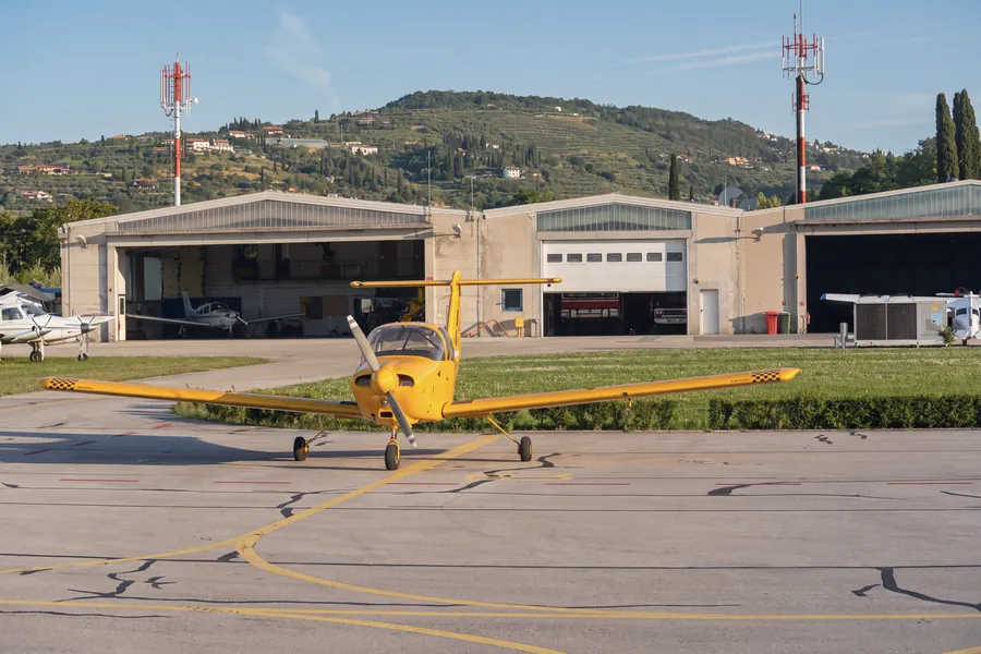 Ein kleines gelbes Propellerflugzeug auf dem Vorfeld des Flughafens Portorož vor den Hangars; links weitere Kleinflugzeuge, zwei rot-weiße Masten, Zypressen und dahinter ein terrassierter Hang mit verstreuten Häusern. Morgenlicht.
