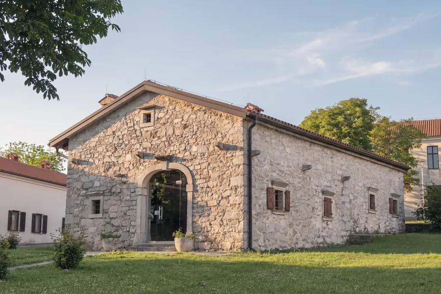 Ein Karst-Steinhaus: Mauern aus sichtbarem Kalkstein, ein steinernes Rundbogenportal mit Glastür, kleine Fenster mit Steinrahmen und Holzläden, Kragsteine an der Fassade, Ziegeldach mit Kamin; davor eine Wiese mit zwei Steinkübeln, seitlich verputzte Gebäude und Bäume. Warmes Licht am späten Nachmittag.