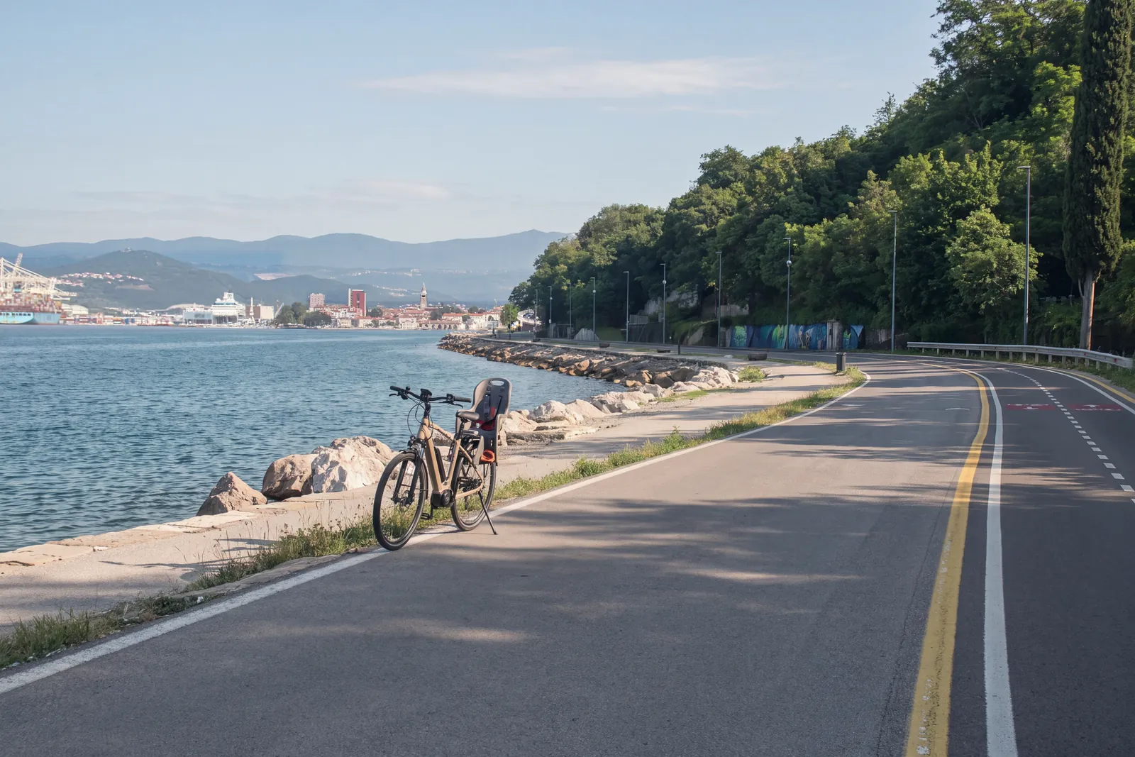 The seaside cycle and footpath between Koper and Izola, on the Parenzana route: an e-bike with a child seat stands on its kickstand at the edge of the path, beside the rock breakwater; across the water, Koper with its bell tower and the port cranes. No people.