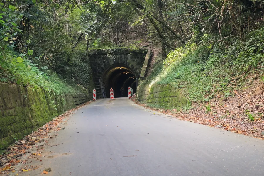 Das Portal des Valeta-Tunnels an der ehemaligen Parenzana-Bahn, heute ein Rad- und Fußweg: ein innen beleuchteter Steinbogen, rot-weiße Poller am Eingang, bemooste Stützmauern aus Stein und Wald zu beiden Seiten, Laub auf dem Asphalt.