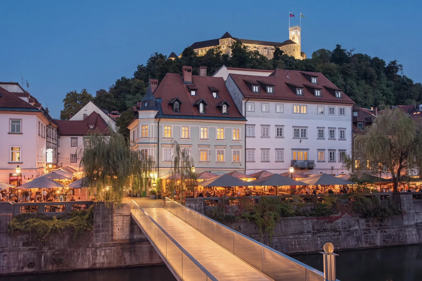 Ljubljana at blue hour: a lit footbridge over the Ljubljanica leads to a riverside terrace of cafés under umbrellas and two old houses with dormer windows; above, on the wooded hill, Ljubljana Castle with its clock tower and two flags.