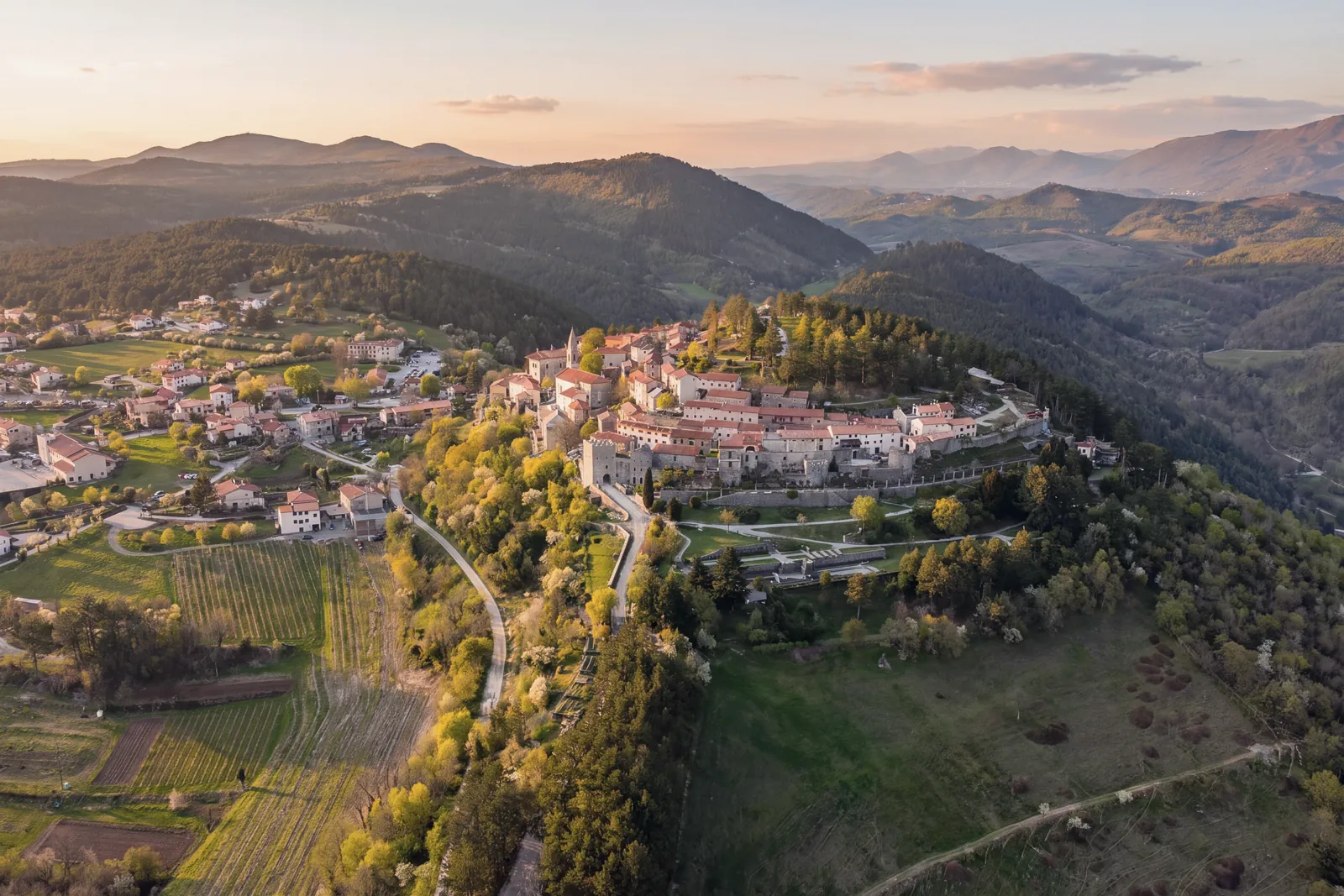 The village of Štanjel on its hill seen from above at sunset: stone houses with tiled roofs, the castle, the walls and the bell tower, terraced gardens below the walls; on the left the newer houses, fields and vineyards; all around, wooded hills and distant mountains.