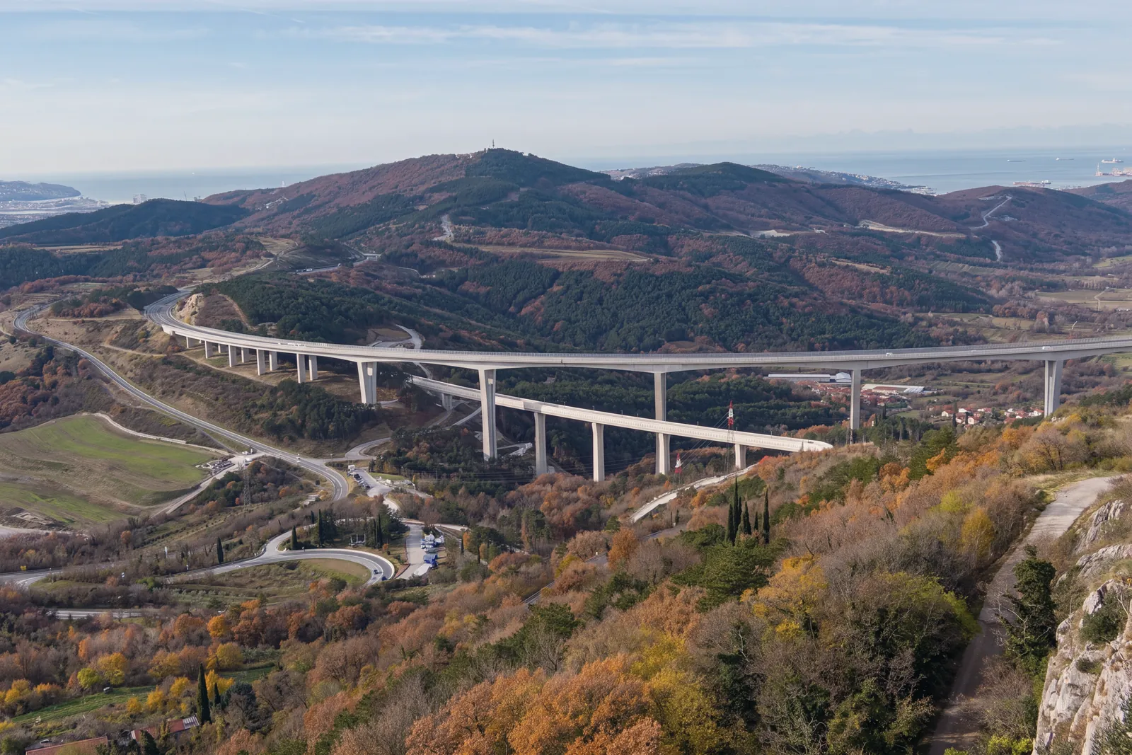 The Črni Kal motorway viaduct seen from high on the Karst edge: a long curve on tall piers, in front a second, lower viaduct, hairpin roads in the valley, wooded hills in autumn colours and the sea in the distance; on the right, the rock of the edge and a path. Side light.