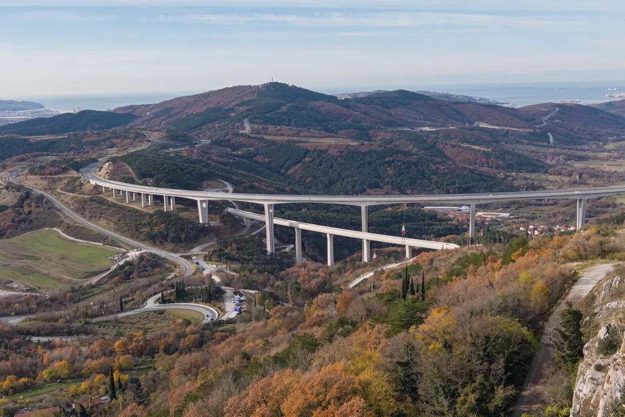 Der Autobahnviadukt von Črni Kal, von oben am Karstrand gesehen: ein langer Bogen auf hohen Pfeilern, davor ein zweiter, niedrigerer Viadukt, Serpentinen im Tal, bewaldete Hügel in Herbstfarben und in der Ferne das Meer; rechts der Fels des Randes und ein Weg. Seitenlicht.