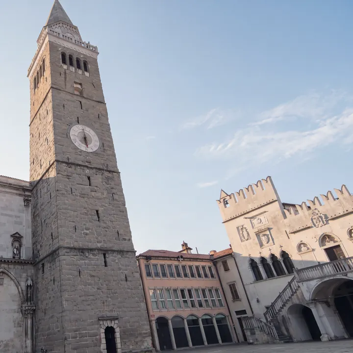 Der Tito-Platz in Koper im ersten Licht, menschenleer: links der steinerne Glockenturm der Kathedrale mit der Uhr, in der Mitte ein rosa Gebäude mit Arkaden, rechts der zinnenbewehrte Prätorenpalast mit Außentreppe und Loggia.
