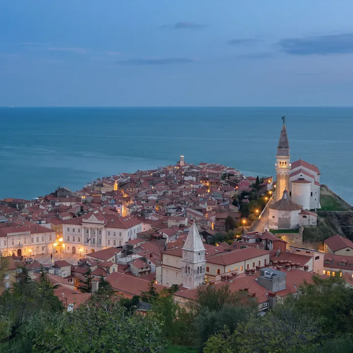 Piran von oben zur blauen Stunde, vom Hügel der Stadtmauer: die roten Dächer der Altstadt bis zur Spitze, der Tartini-Platz mit dem Rathaus und den ersten brennenden Laternen, rechts die Kirche St. Georg mit beleuchtetem Glockenturm und dem achteckigen Baptisterium, im Vordergrund ein weiterer Glockenturm; dahinter das offene Meer.