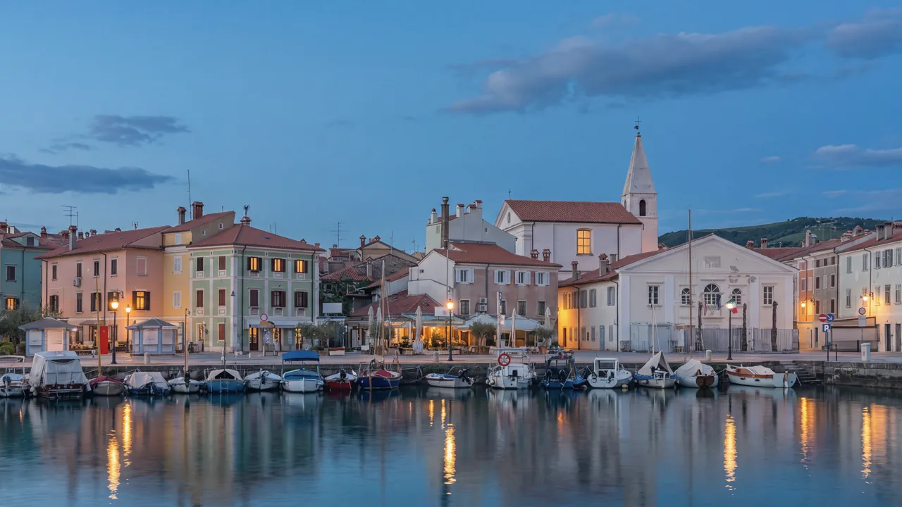 Der kleine Hafen von Izola zur blauen Stunde: Fischer- und Freizeitboote an der Kaimauer, Häuser im venezianischen Stil in Pastellfarben, eine Kirche mit Glockenturm und ein weißes klassizistisches Gebäude; Laternen und einige Fenster erleuchtet, Spiegelungen auf dem ruhigen Wasser.