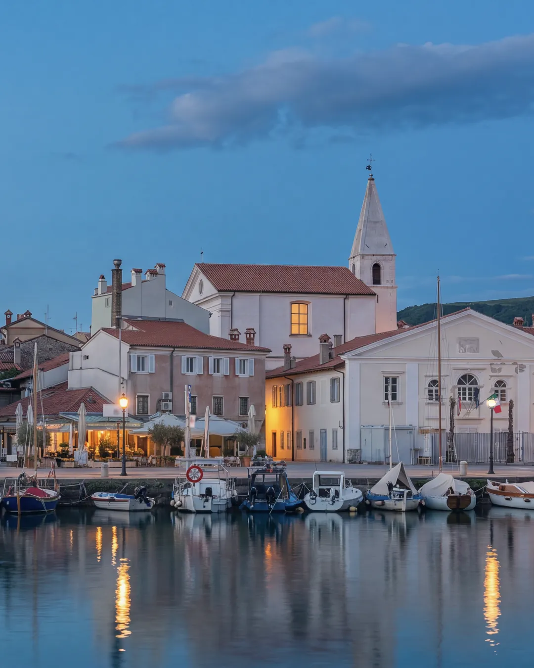 Izola's small harbour at blue hour: fishing and pleasure boats moored along the quay, Venetian-style houses in pastel colours, a church with its bell tower and a white classical building; street lamps and some windows lit, reflections on the calm water.