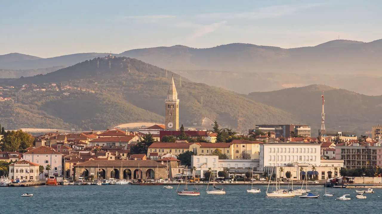 Koper über das Wasser von Žusterna aus gesehen, zur goldenen Stunde: der Glockenturm der Kathedrale, die roten Dächer der Altstadt, ein Steinbau mit Arkaden an der Uferpromenade, die weißen modernen Gebäude am Ufer, ein rot-weißer Gittermast und ein Kran rechts, davor ankernde Segelboote; dahinter bewaldete Hügel.
