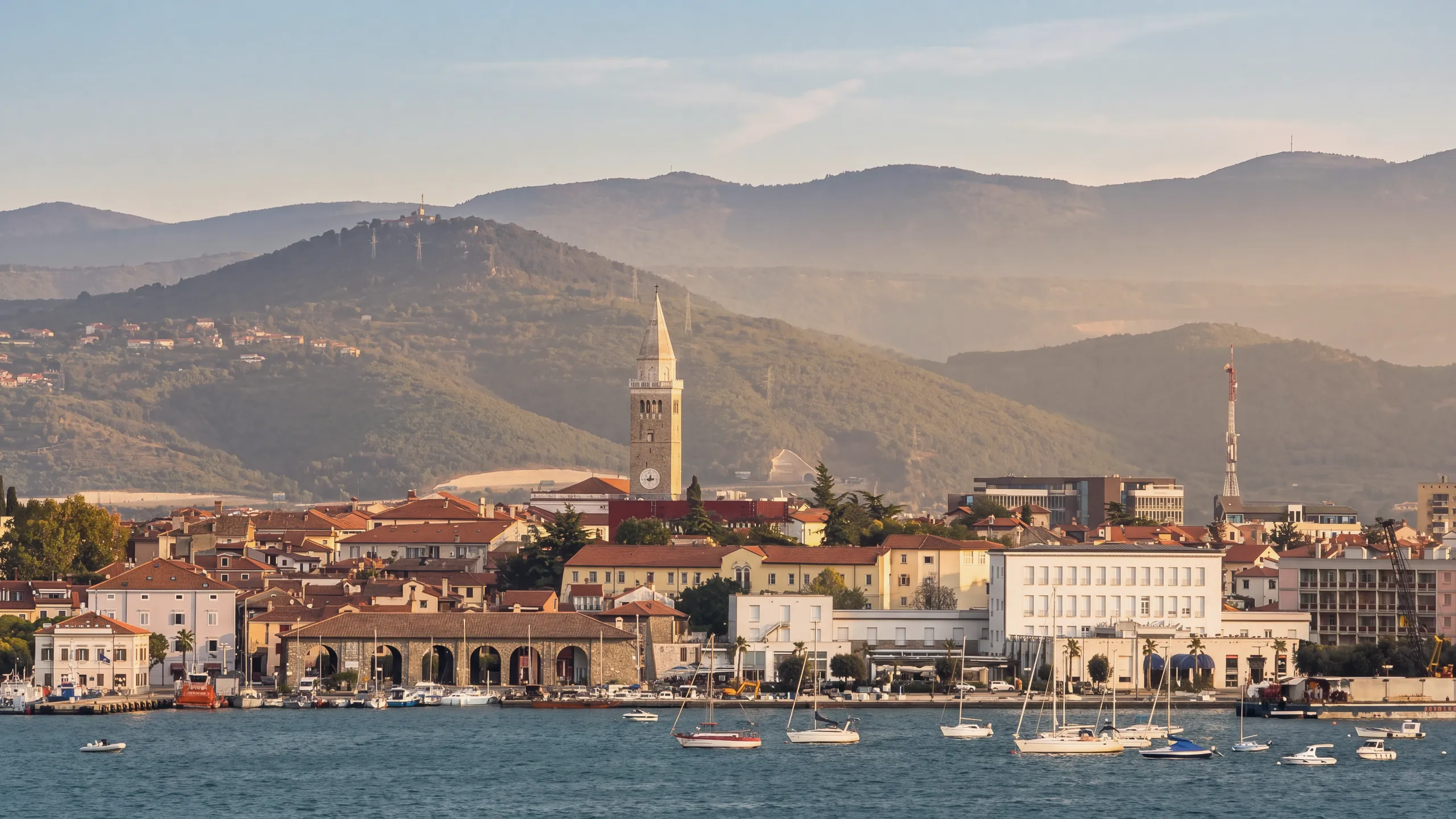 Koper seen across the water from Žusterna at golden hour: the cathedral bell tower, the red roofs of the old town, a stone building with arcades on the waterfront, the white modern buildings on the shore, a red-and-white lattice mast and a crane on the right, sailing boats moored in front; behind, wooded hills.