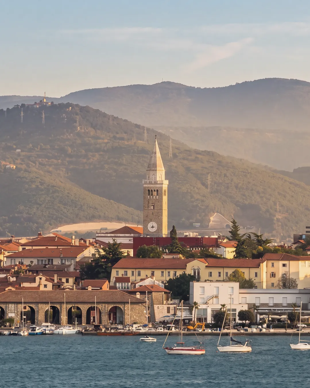 Koper seen across the water from Žusterna at golden hour: the cathedral bell tower, the red roofs of the old town, a stone building with arcades on the waterfront, the white modern buildings on the shore, a red-and-white lattice mast and a crane on the right, sailing boats moored in front; behind, wooded hills.