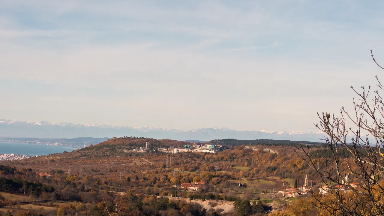 Der Karst um Črnotiče im Herbst: braune und goldene Wälder, ein Hügel mit einem Steinbruch und seinen Anlagen, Häuser und ein Glockenturm rechts, Strommasten; links das Meer des Golfs von Triest mit einer Stadt an der Küste, am Horizont die verschneiten Alpen; kahle Zweige rechts im Vordergrund. Licht am späten Nachmittag.