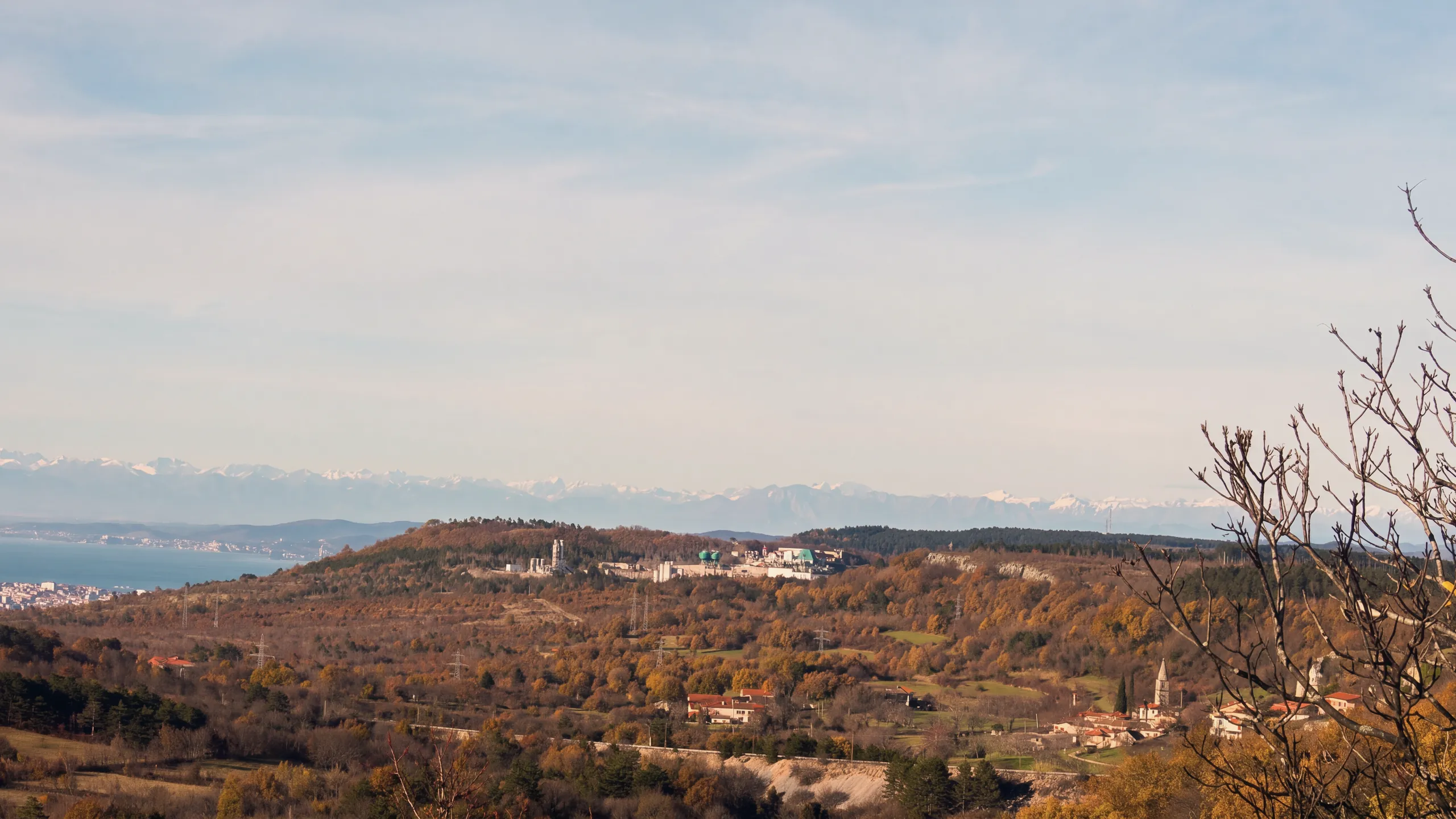 The Karst around Črnotiče in autumn: brown and golden woods, a hill with a quarry and its plant, houses and a bell tower on the right, power pylons; on the left the sea of the Gulf of Trieste with a town on the coast, snow-capped Alps on the horizon; bare branches in the right foreground. Late-afternoon light.