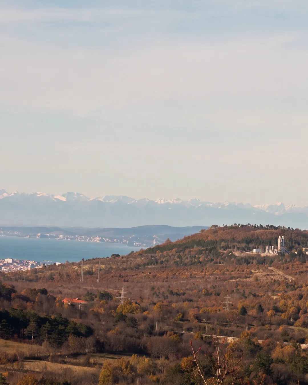 The Karst around Črnotiče in autumn: brown and golden woods, a hill with a quarry and its plant, houses and a bell tower on the right, power pylons; on the left the sea of the Gulf of Trieste with a town on the coast, snow-capped Alps on the horizon; bare branches in the right foreground. Late-afternoon light.