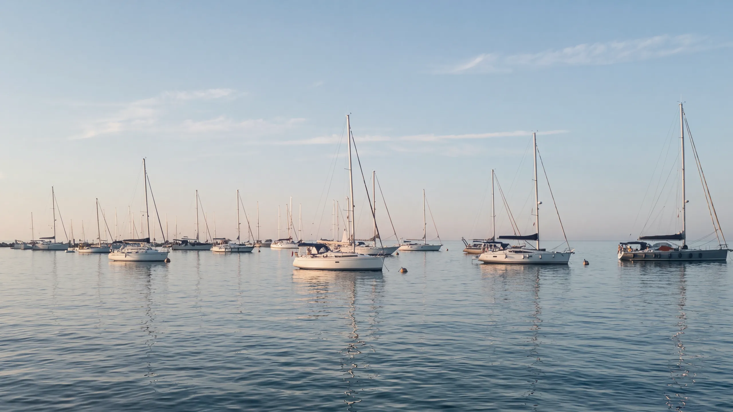 Sailing boats moored on buoys in the bay of Portorož, in the morning: white hulls, masts and rigging lit by the low sun from behind the camera, calm sea with reflections; on the left the breakwater with its light, the open sea on the horizon.