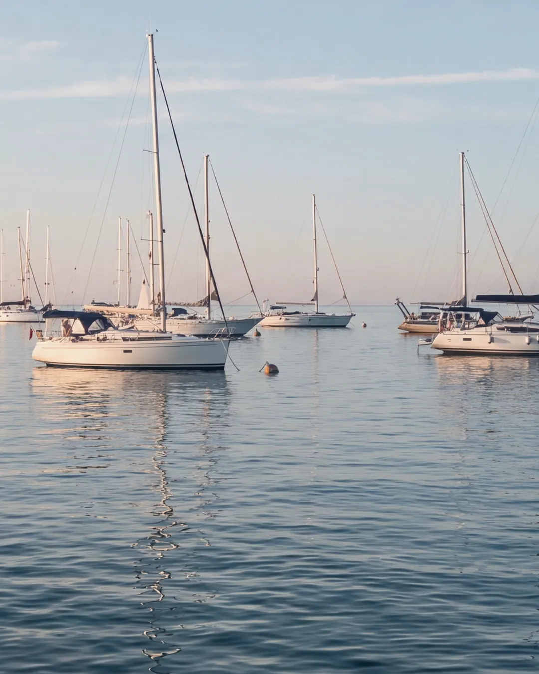 Segelboote an Bojen in der Bucht von Portorož am Morgen: weiße Rümpfe, Masten und Wanten im Licht der tiefen Sonne im Rücken, ruhiges Meer mit Spiegelungen; links der Wellenbrecher mit seinem Leuchtfeuer, am Horizont das offene Meer.